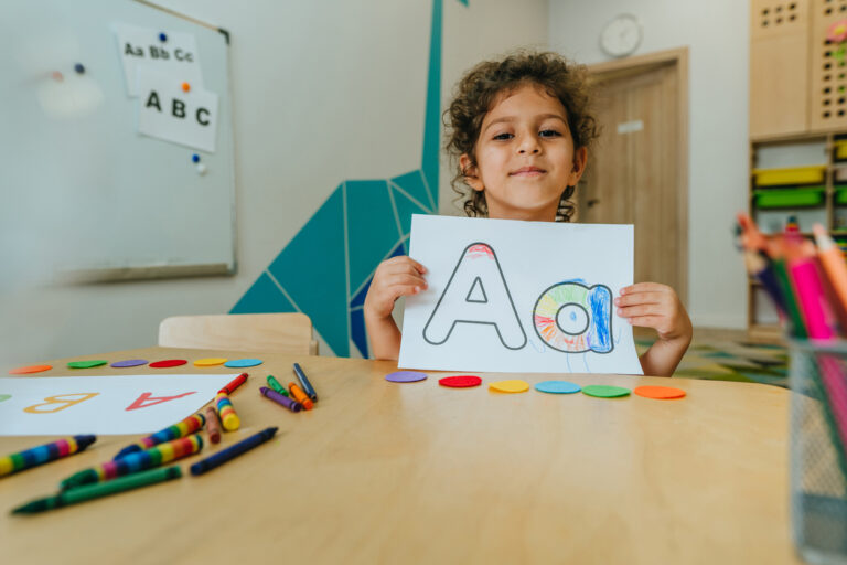 Kindergarten aged girl in a classroom who is holding up a drawing